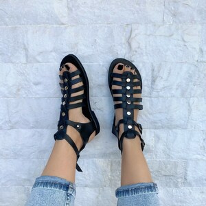 Puede incluir: Sandalias gladiadoras de cuero negro con hebillas y tachuelas doradas. Las sandalias tienen una correa que va alrededor del tobillo y un cierre de hebilla.