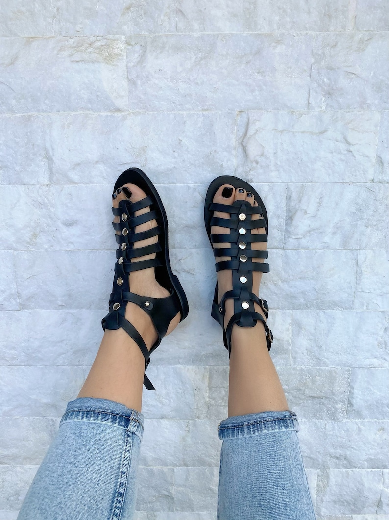 Puede incluir: Sandalias gladiadoras de cuero negro con hebillas y tachuelas doradas. Las sandalias tienen una correa que va alrededor del tobillo y un cierre de hebilla.