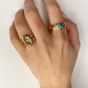 May include: Two gold rings on a hand. One ring has a turquoise stone and the other ring has an eye design.