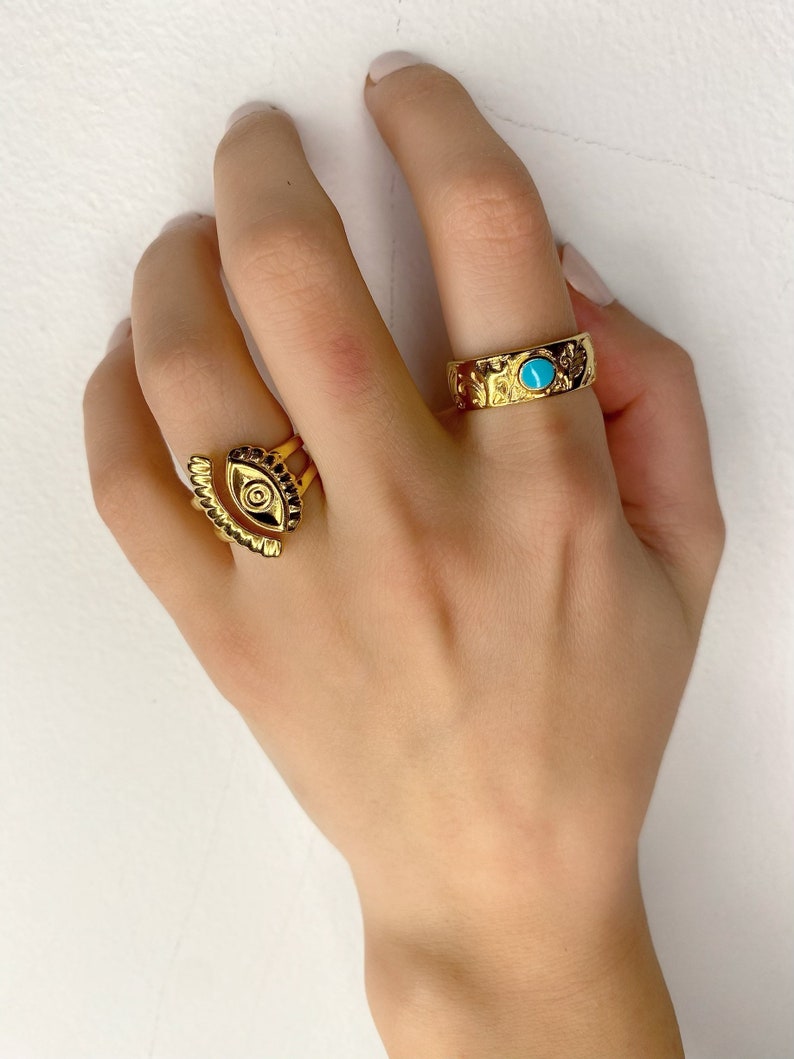 May include: Two gold rings on a hand. One ring has a turquoise stone and the other ring has an eye design.
