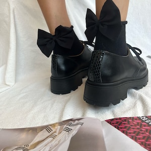 May include: Black platform shoes with black bow socks. The shoes have a chunky heel and a rounded toe. The socks have a large black bow on the top.