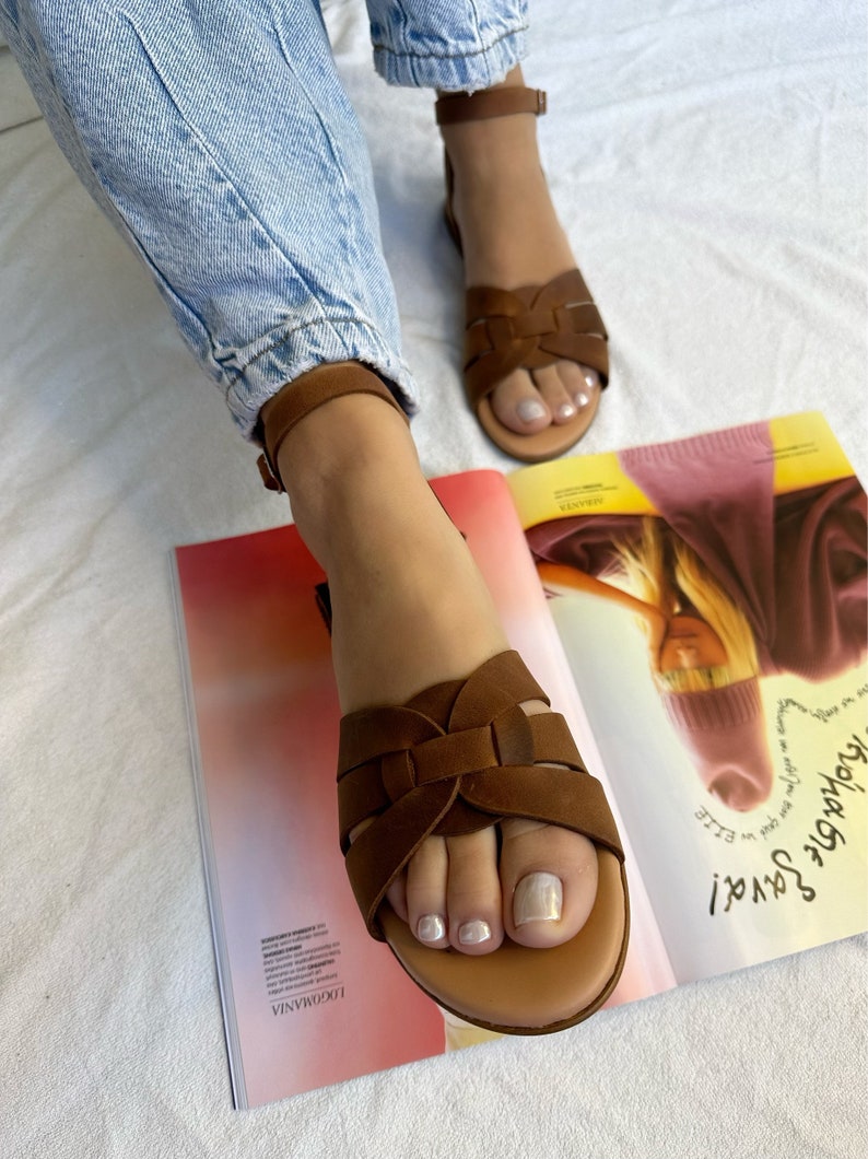 Puede incluir: Sandalias de cuero marr&oacute;n con un dise&ntilde;o de tiras tejidas. Las sandalias tienen una tira para el tobillo con cierre de hebilla. Las sandalias se llevan en los pies de una persona.