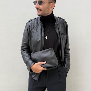 Op de afbeelding: Een zwart leren jack met ritssluiting, een zwarte coltrui, een zwarte broek met een dunne riem en een zwarte leren clutch. De outfit bevat een zilveren kruishanger aan een zwart koord en een zonnebril met donkere glazen.