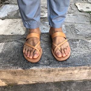 May include: A pair of tan leather sandals with criss-cross straps. The sandals are worn on a person's feet.