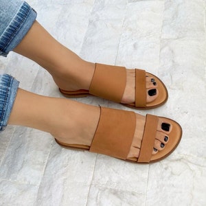 May include: A pair of brown leather sandals with two straps across the top of the foot. The sandals have a flat sole and are worn by a person with bare feet.