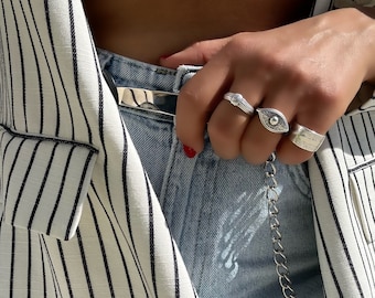 Puede incluir: Primer plano de una mano con tres anillos de plata. Los anillos son una banda simple, un anillo en forma de ojo y un anillo con un diseño texturizado. La persona lleva una camisa blanca, una chaqueta de rayas y pantalones vaqueros azules.