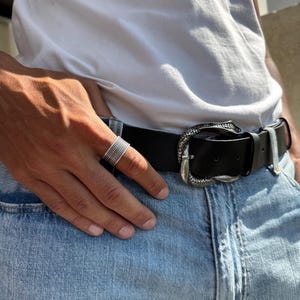 May include: A silver ring with multiple bands is worn on a hand. A black leather belt with a decorative silver buckle is fastened around the waist. The person is wearing a white t-shirt and blue jeans.