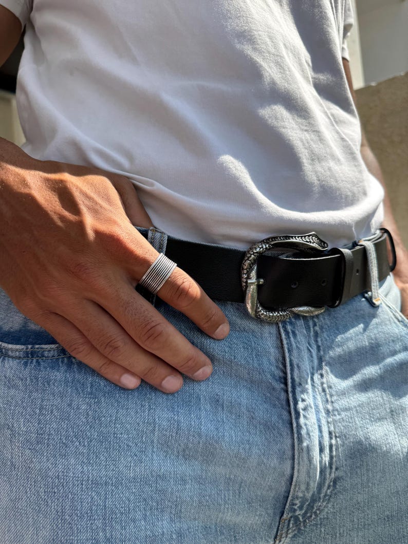 May include: A silver ring with multiple bands is worn on a hand. A black leather belt with a decorative silver buckle is fastened around the waist. The person is wearing a white t-shirt and blue jeans.
