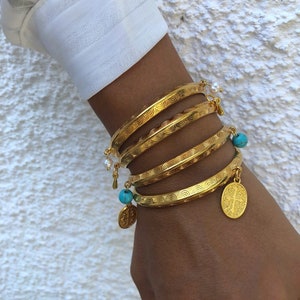 Pode incluir: Um conjunto de cinco pulseiras douradas com superf&iacute;cie texturizada. As pulseiras est&atilde;o empilhadas no pulso de uma pessoa e apresentam pequenos pingentes turquesa e p&eacute;rola.