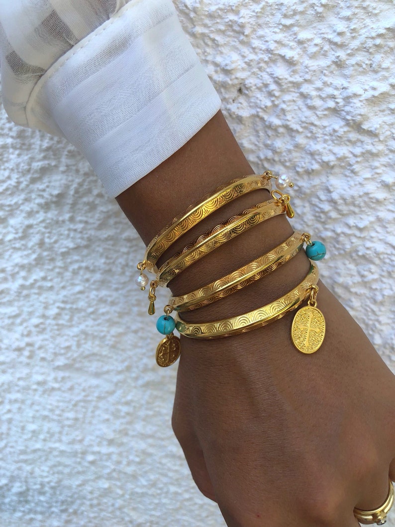 Pode incluir: Um conjunto de cinco pulseiras douradas com superf&iacute;cie texturizada. As pulseiras est&atilde;o empilhadas no pulso de uma pessoa e apresentam pequenos pingentes turquesa e p&eacute;rola.