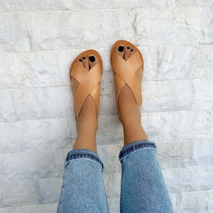 Puede incluir: Sandalias de cuero marr&oacute;n claro con tiras cruzadas. Las sandalias se llevan en los pies de una persona con pantalones vaqueros azules.