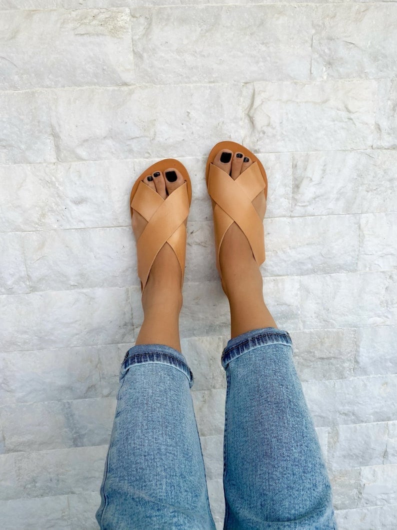 Puede incluir: Sandalias de cuero marr&oacute;n claro con tiras cruzadas. Las sandalias se llevan en los pies de una persona con pantalones vaqueros azules.