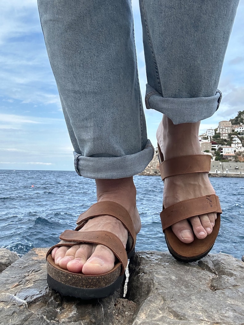 May include: Brown leather sandals with adjustable straps and cork footbeds. The sandals have a thick, black sole. The person is wearing light blue jeans with the cuffs rolled up. The background shows a body of water and a cloudy sky.