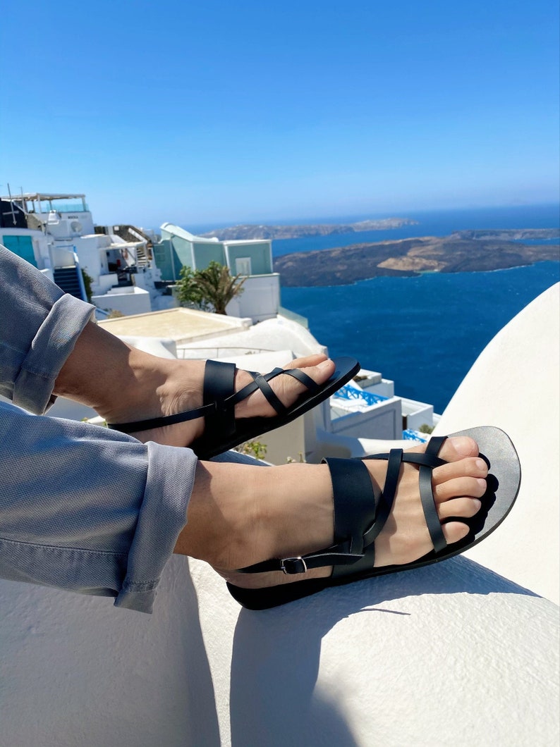May include: A pair of black leather sandals with a criss-cross strap design. The sandals are worn by a person sitting on a white wall overlooking a blue ocean.