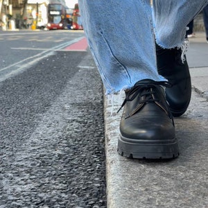 May include: A pair of black leather lace-up shoes with a thick sole. The shoes are worn by a person wearing blue jeans.