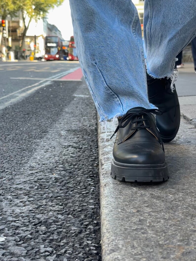 May include: A pair of black leather lace-up shoes with a thick sole. The shoes are worn by a person wearing blue jeans.