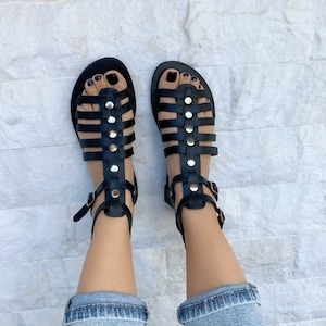 Puede incluir: Sandalias gladiadoras de cuero negro con tachuelas y hebillas doradas. Las sandalias tienen un dise&ntilde;o enjaulado y se usan en los pies.