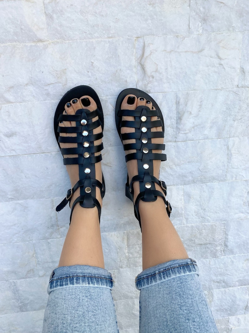 Puede incluir: Sandalias gladiadoras de cuero negro con tachuelas y hebillas doradas. Las sandalias tienen un dise&ntilde;o enjaulado y se usan en los pies.