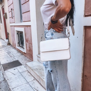 Puede incluir: Un bolso bandolera de cuero blanco con cierre de solapa y cadena plateada. El bolso lo lleva una persona que lleva pantalones vaqueros de color azul claro y una camiseta blanca.