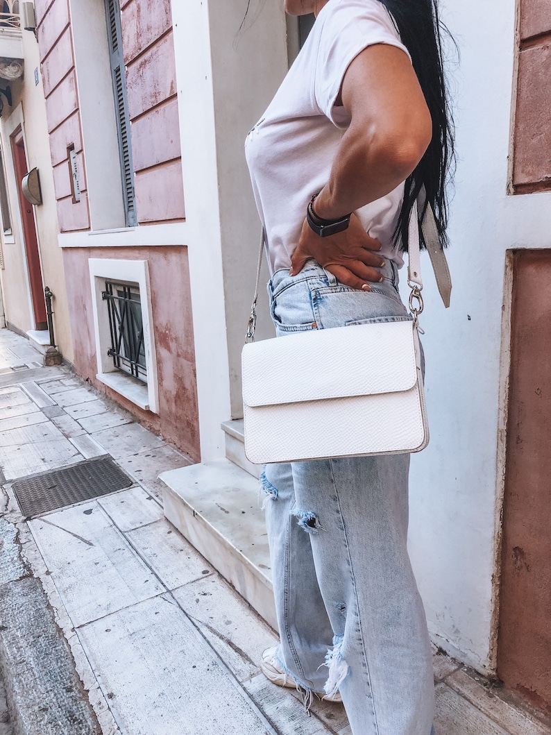 Puede incluir: Un bolso bandolera de cuero blanco con cierre de solapa y cadena plateada. El bolso lo lleva una persona que lleva pantalones vaqueros de color azul claro y una camiseta blanca.