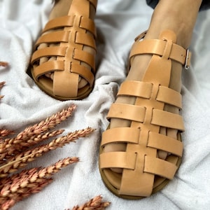 K&ouml;nnte beinhalten: Tanfarbene Ledersandalen mit geflochtenem Design und Schnallenverschluss. Die Sandalen befinden sich auf einem wei&szlig;en Hintergrund.