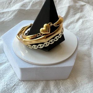 May include: A set of three gold bracelets with a heart charm. The bracelets are stacked on a black pyramid-shaped stand.