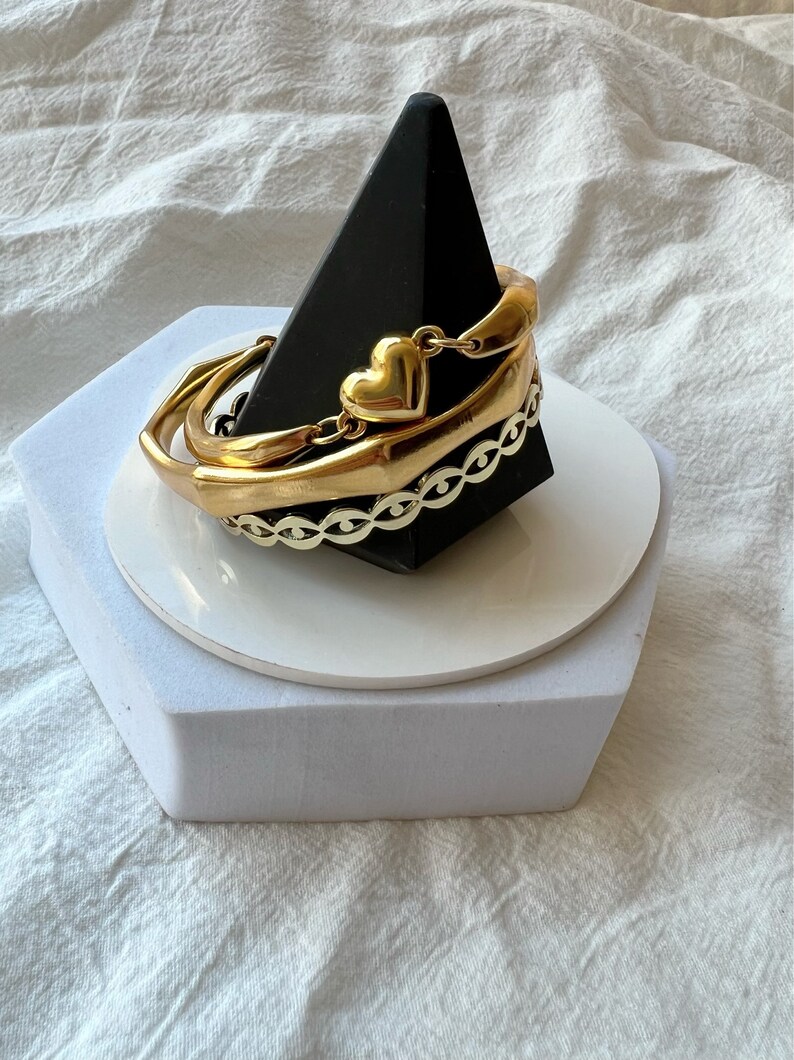 May include: A set of three gold bracelets with a heart charm. The bracelets are stacked on a black pyramid-shaped stand.