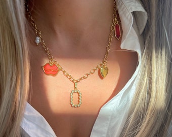 Pode incluir: Um colar de corrente de ouro com vários pingentes, incluindo um lábio vermelho, um coração, uma pérola e uma letra O com strass.