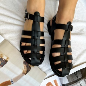 Op de afbeelding: Zwarte leren sandalen met een kooi-ontwerp en een gespsluiting. De sandalen hebben een platte zool en zijn gemaakt van geweven leer.