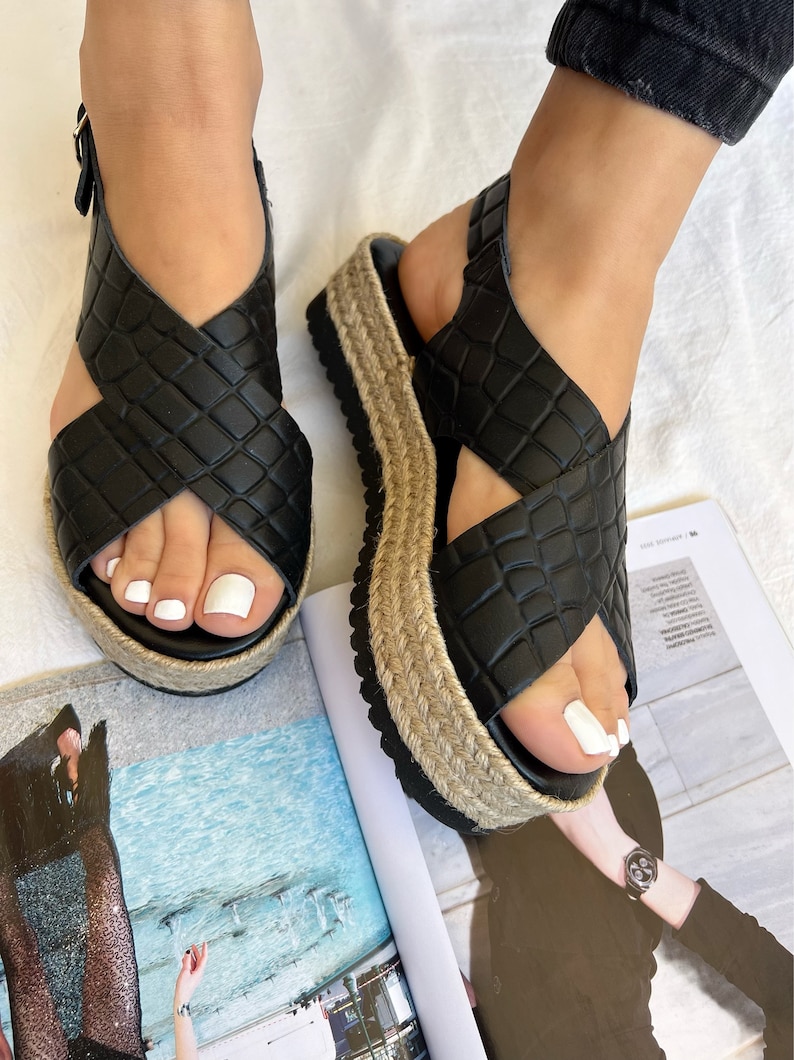 May include: Black platform sandals with a crisscross design. The sandals have a textured, faux-leather upper and a woven jute-style platform sole. The ankle strap features a buckle closure. The sandals are shown on a white surface.
