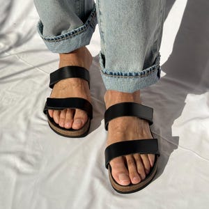 May include: Black leather sandals with two straps and a cork footbed. The sandals are worn with light blue denim jeans with rolled-up cuffs. The sandals are shown on a white background.