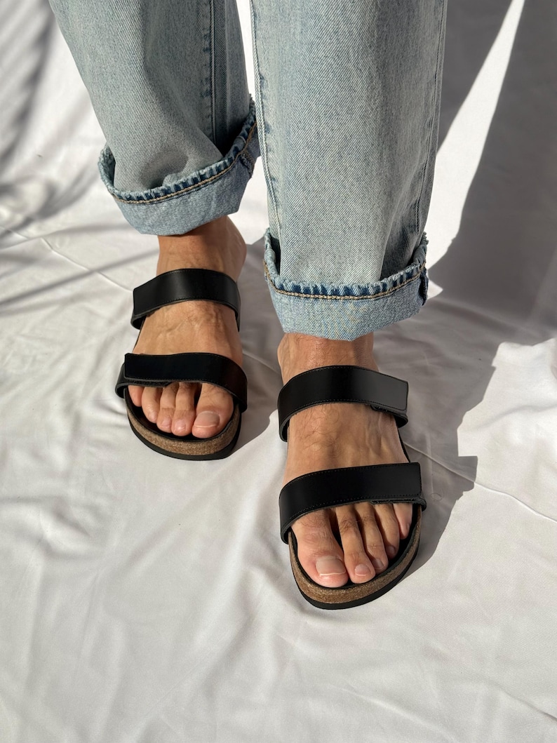 May include: Black leather sandals with two straps and a cork footbed. The sandals are worn with light blue denim jeans with rolled-up cuffs. The sandals are shown on a white background.