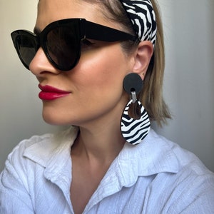 Könnte beinhalten: Eine Frau trägt eine schwarze Cat-Eye-Sonnenbrille, ein weißes Hemd und ein schwarz-weißes Zebra-Stirnband. Sie trägt auch große schwarz-weiße Zebra-Ohrringe mit einem schwarzen Kreis-Akzent.