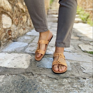 Puede incluir: Sandalias de cuero color canela con detalle de hebilla y correa. Las sandalias tienen suela plana y se llevan en los pies.