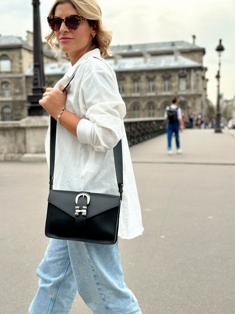 Puede incluir: Una mujer con una camisa blanca y pantalones vaqueros azules lleva un bolso bandolera de cuero negro con una hebilla plateada. El bolso tiene una correa larga y un cierre de solapa.