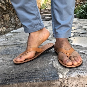 Pode incluir: Um par de chinelos de couro castanho claro com sola preta. As sand&aacute;lias t&ecirc;m um design simples de tanga e s&atilde;o usadas numa superf&iacute;cie de pedra. A imagem mostra as sand&aacute;lias nos p&eacute;s de uma pessoa.