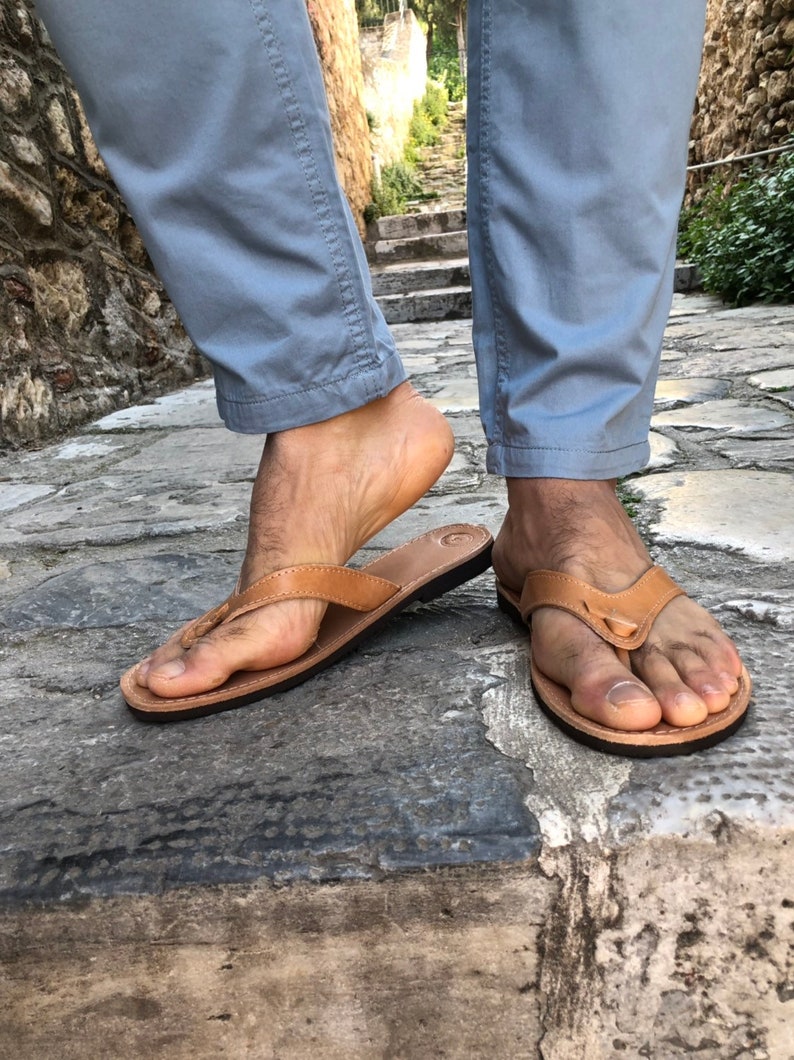 Pode incluir: Um par de chinelos de couro castanho claro com sola preta. As sand&aacute;lias t&ecirc;m um design simples de tanga e s&atilde;o usadas numa superf&iacute;cie de pedra. A imagem mostra as sand&aacute;lias nos p&eacute;s de uma pessoa.