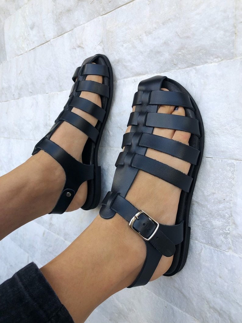 May include: Black leather sandals with a woven strap design and a buckle closure. The sandals have a flat sole and are made of high-quality leather.