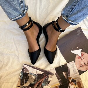 May include: Black flats with pointed toes and ankle straps adorned with gold-colored studs. The shoes are paired with rolled-up blue jeans. Fashion magazines are scattered around the shoes.
