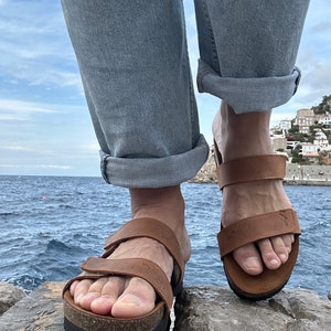 May include: Brown leather sandals with adjustable straps and cork footbeds. The sandals have a thick, black sole. The person is wearing light blue jeans with the cuffs rolled up. The background shows a body of water and a cloudy sky.