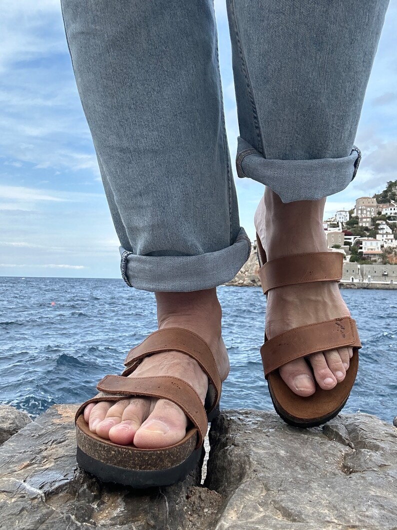 May include: Brown leather sandals with adjustable straps and cork footbeds. The sandals have a thick, black sole. The person is wearing light blue jeans with the cuffs rolled up. The background shows a body of water and a cloudy sky.
