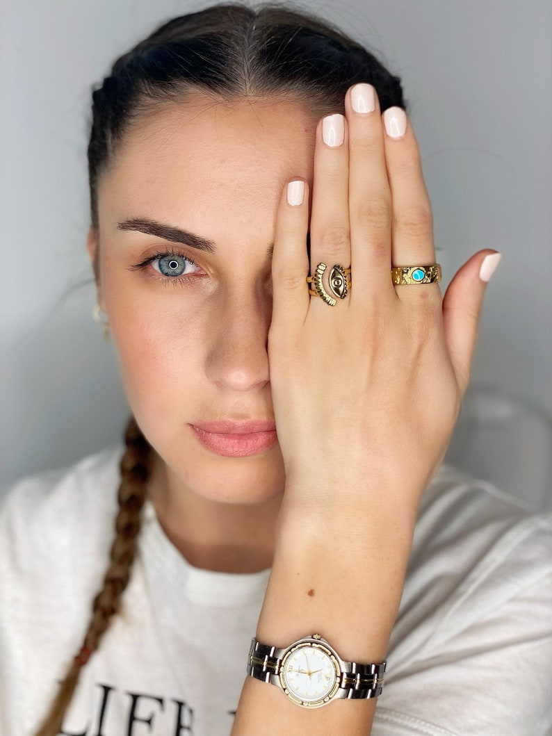 May include: A woman wearing a white shirt with the word "LIFE" printed on it, with a gold watch on her wrist. She is wearing three gold rings on her finger, one with a turquoise stone and another with an eye design.