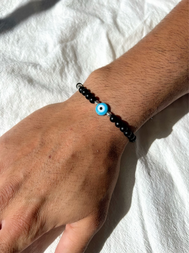 May include: A black beaded bracelet with a blue evil eye charm. The bracelet is on a person's wrist.
