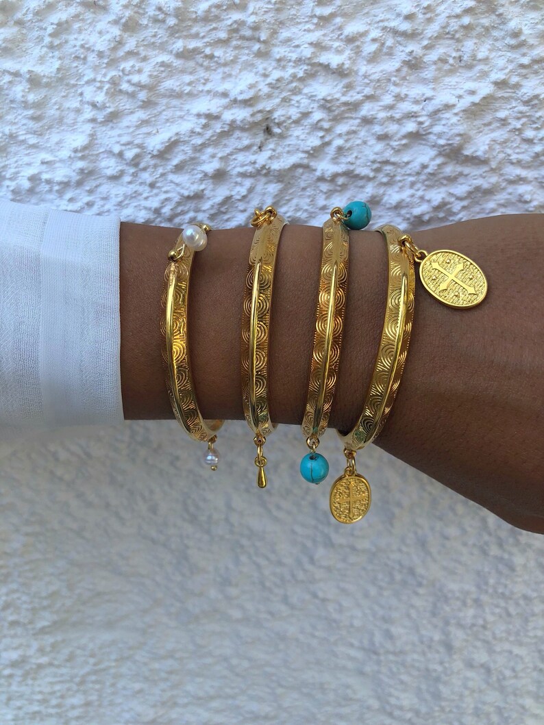 Pode incluir: Cinco pulseiras de ouro com padr&otilde;es decorativos e pingentes. Os pingentes incluem uma p&eacute;rola, uma conta turquesa, um pingente em forma de l&aacute;grima e uma moeda de ouro com um desenho de cruz.