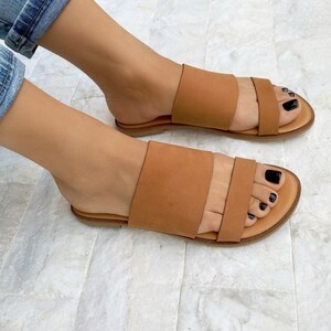 May include: A pair of brown leather sandals with two straps across the top of the foot. The sandals have a flat sole and are worn on a person's feet.