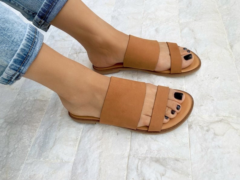 May include: A pair of brown leather sandals with two straps across the top of the foot. The sandals have a flat sole and are worn on a person's feet.