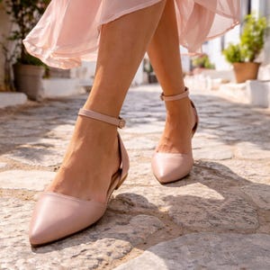 May include: Pale pink flats with ankle straps. The shoes have a pointed toe and a low heel. The image shows the shoes being worn while walking on a cobblestone street. The person is wearing a pale pink dress.