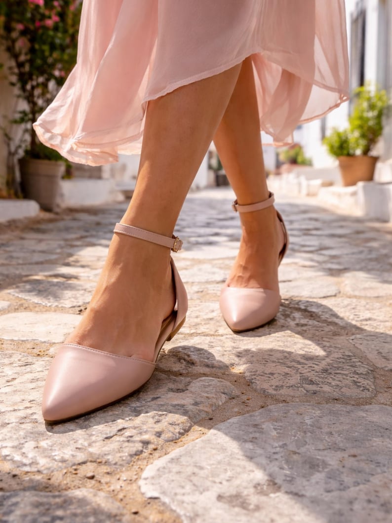 May include: Pale pink flats with ankle straps. The shoes have a pointed toe and a low heel. The image shows the shoes being worn while walking on a cobblestone street. The person is wearing a pale pink dress.