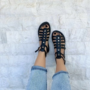 Puede incluir: Sandalias gladiadoras de cuero negro con detalles dorados y hebillas. Las sandalias se llevan puestas en los pies de una persona con pantalones vaqueros azules.