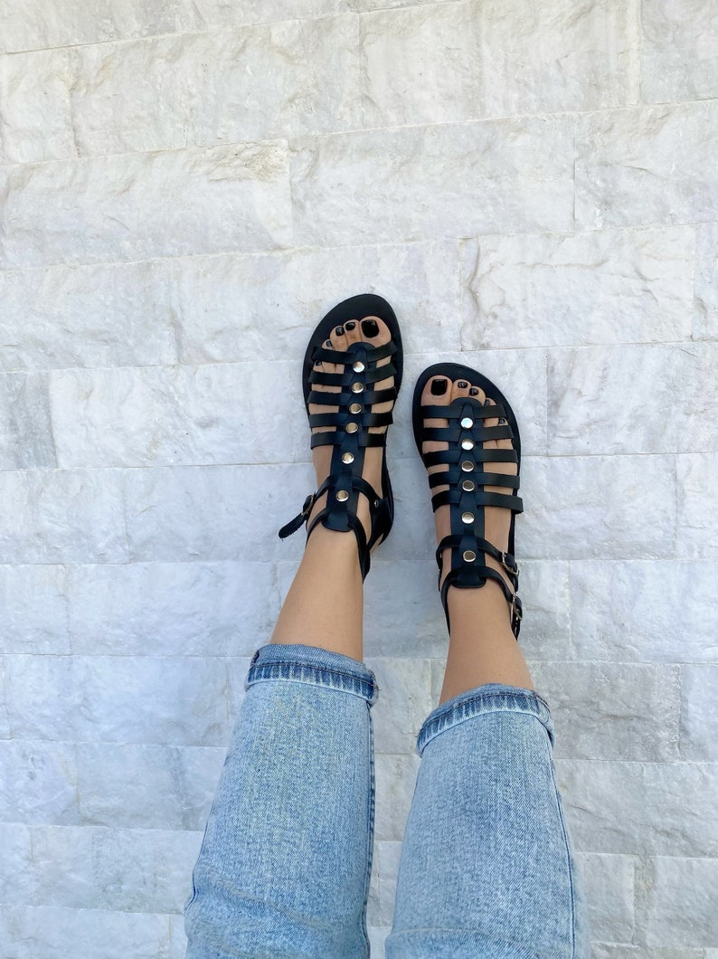 Puede incluir: Sandalias gladiadoras de cuero negro con detalles dorados y hebillas. Las sandalias se llevan puestas en los pies de una persona con pantalones vaqueros azules.